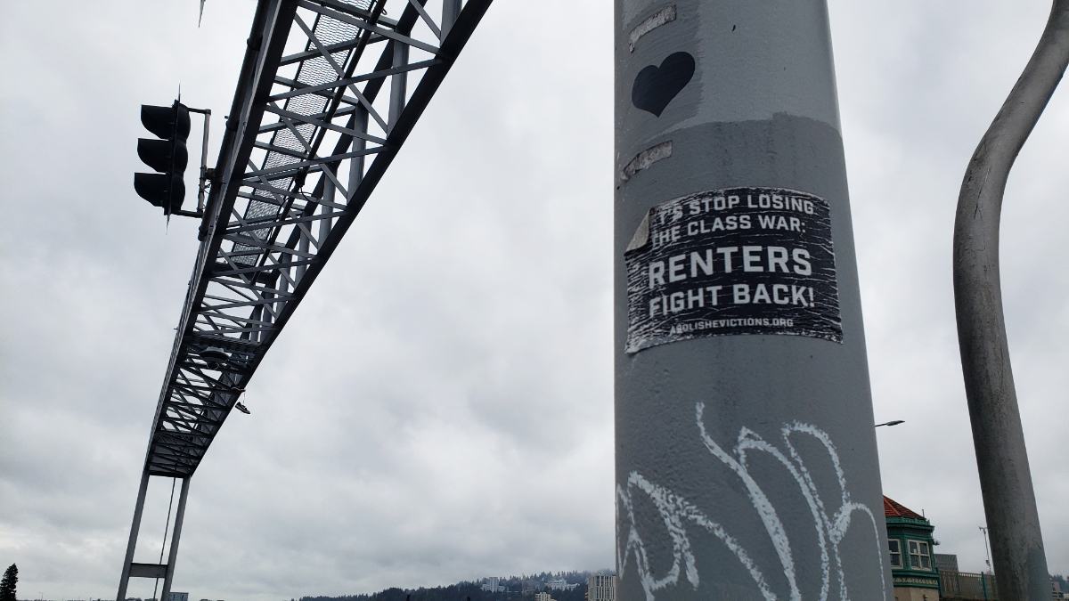 a concrete pole next to an overhead road sign with a sticker saying 'Let's stop losing the class war: Renters Fight Back! abolishevictions.org