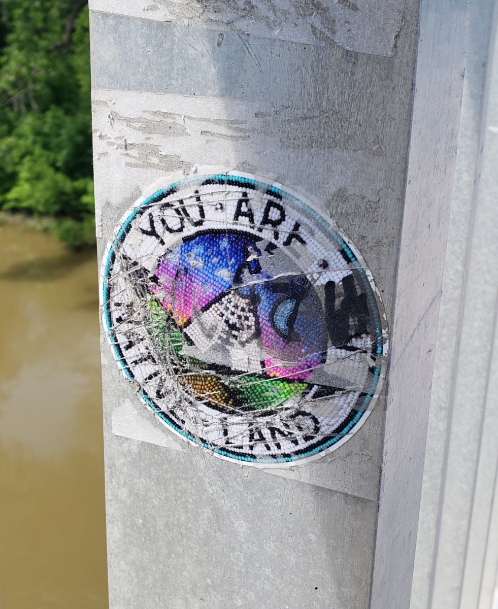 A railing on a bridge over brown water and a bank with green trees with a round sticker saying 'YOU ARE ON NATIVE LAND' around a picture of a tepee against a blue and purple sky between green hills, made to look like it's made with beads