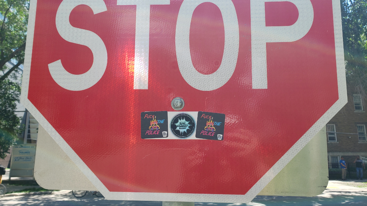 the bottom 60% of a stop sign with three stickers right below the bolt keeping it to the post: two edge stickers that have a Lite Brite style image of a cop car on fire and the words 'Fuck the Police', and one in the middle for Winnipeg Critical Mass.