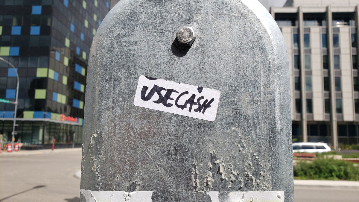 a silver lamppost with a sticker on a panel cover with 'USE CASH' written in black marker, and office buildings across the street in the background.