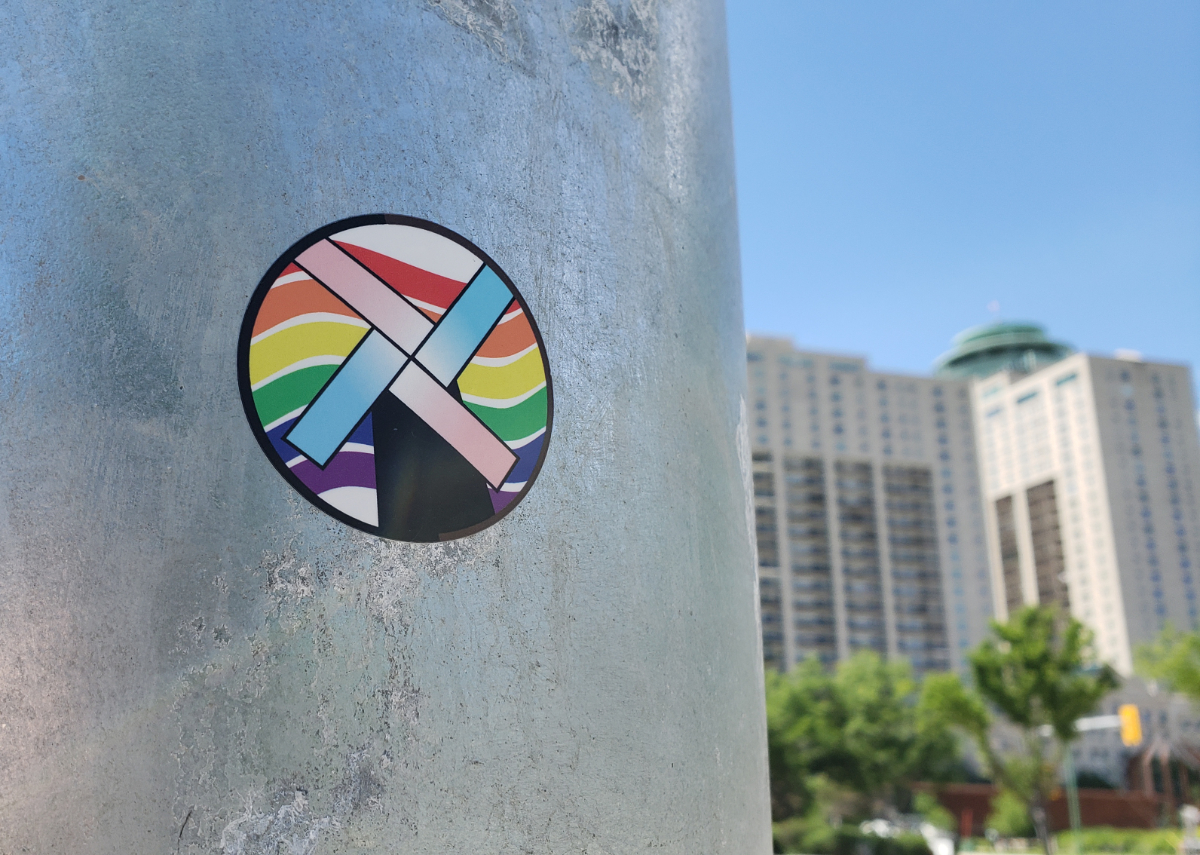 a silver pole with a round sticker on it that shows the pride rainbow colours as waves in the background and a windmill with the trans flag colours as blades