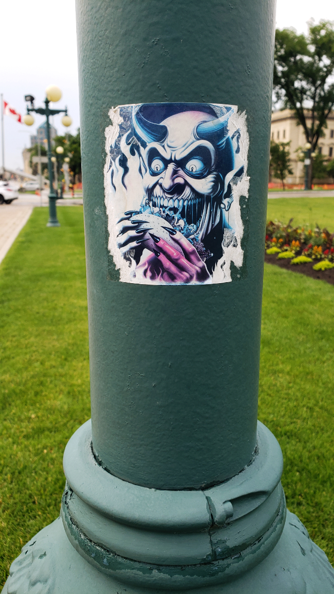 a green lamppost with a high contrast sticker with teal and magenta tones of a devil eating a taco. the background features a lawn with a garden, another lamp post, a Canadian flag, and some distant buildings.