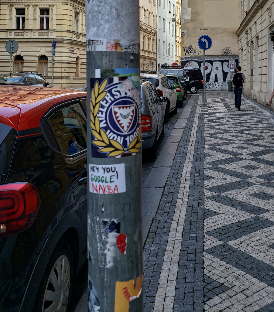 a metal pole next to parked cars with various stickers on it, one of which says 'hey you! google Nakba!' in Palestinian colours