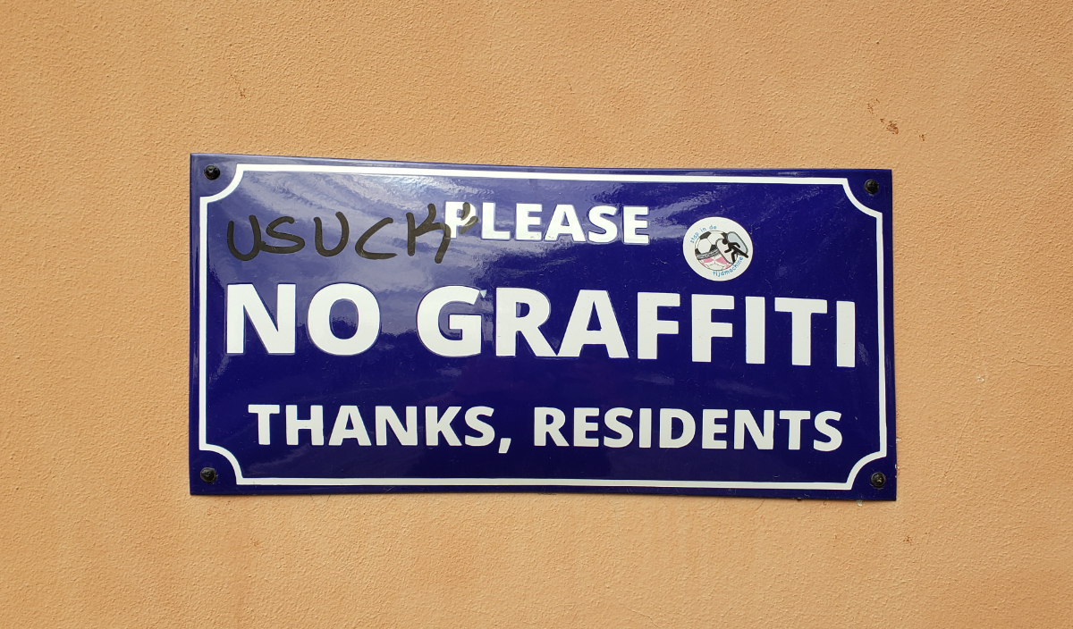 a beigey orange exterior wall with a dark blue sign with white lettering saying 'Please, No Graffiti, Thanks, Residents' and a sticker on the top righrt of that plus black permanent marker on the top left saying 'U SUCK'