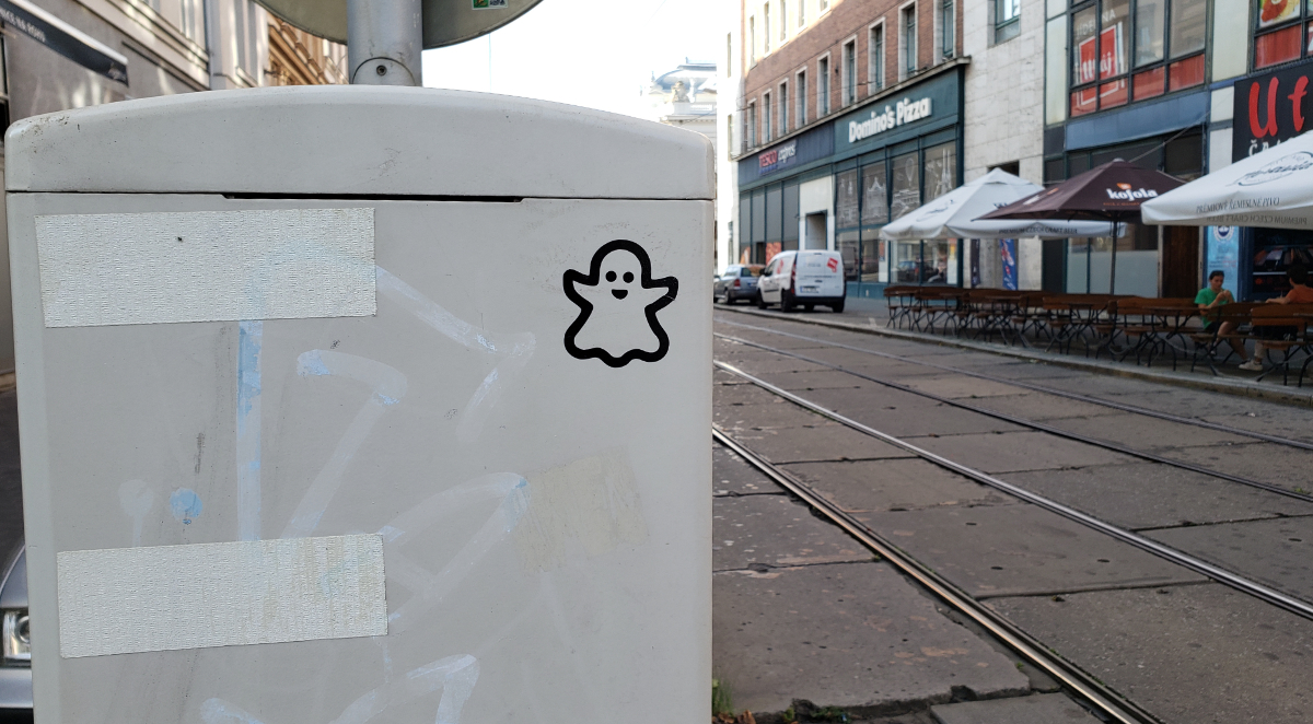 a grey power box with a lil cute ghost sticker on it. to the right is a road with tram tracks and a sidewalk with patio seating for various places, including, naturally, a Domino's restaurant