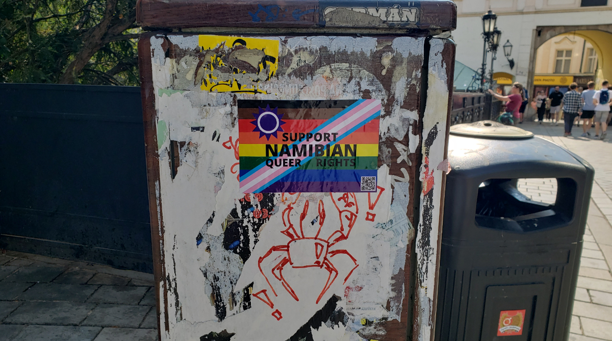 A trash bin next to a walkway towards an arch with some generic graffiti (including a crab drawn in red marker) plus a pride flag sticker saying 'Support Namibian Queer Rights'