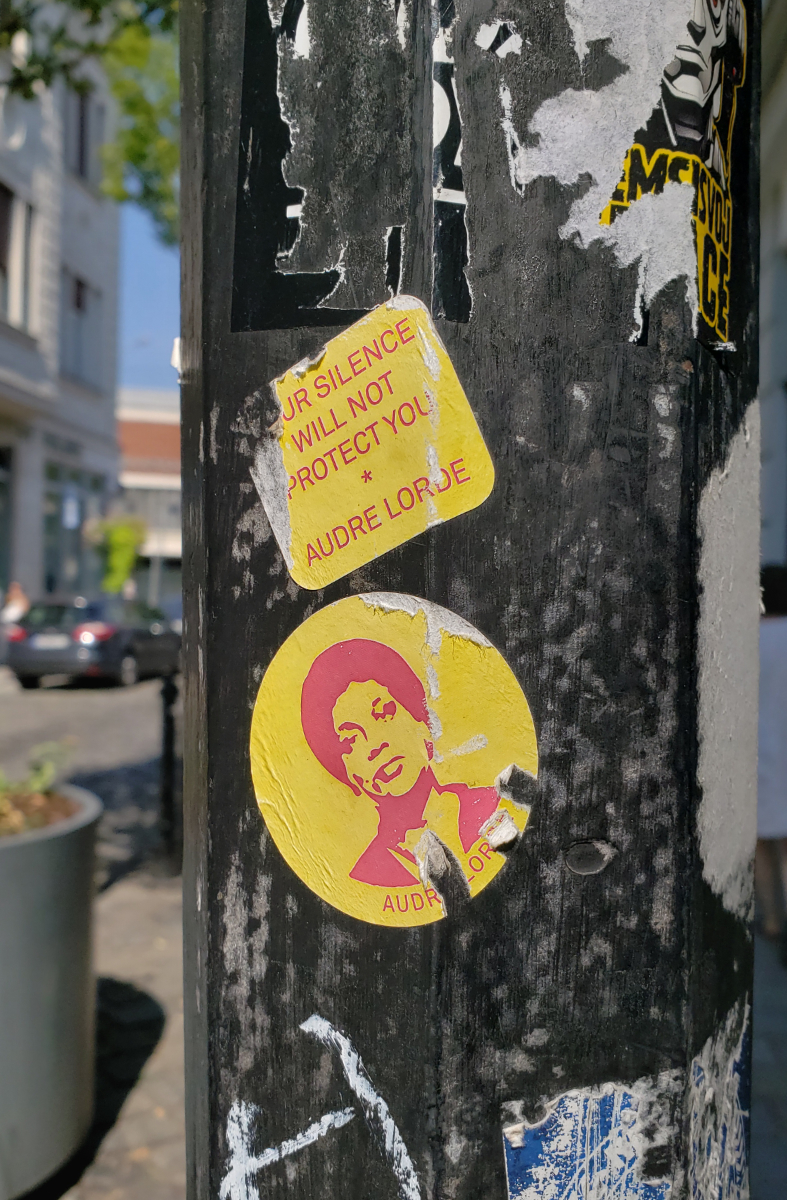 a black lamppost with many peeled off stickers, and two yellow stickers: one partially torn off yellow rectangle with text one can fill in the gaps to say 'YOUR SILENCE WILL NOT PROTECT YOU * AUDRE LORDE' and a yellow circle below with an image of Audre Lorde.