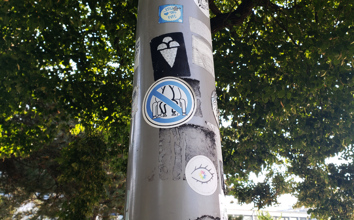 a grey pole against a background of lush green trees, with stickers on the pole including: black background with silver shapes looking like a three-scoop ice cream cone; a drawing of manspreading with a blue 'no' circle around it; an eye with a white pupil and rainbow tendrils around it