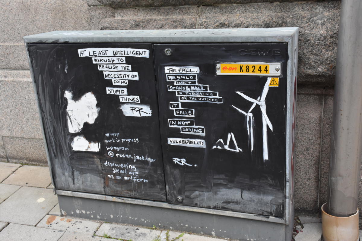 Malmo, Sweden a black power box with white paint saying various messages: 'at least intelligent enough to realize the necessity of doing stupid things'; '#WIP work in progress, (nearly illegible instagram username) discovering street art as an artform'; 'the fall. the world doesn't stand & fall in the eroding or the outcome. it falls in not daring. vulnerability' next to a drawing of one collapsed wind turbine next to two still standing