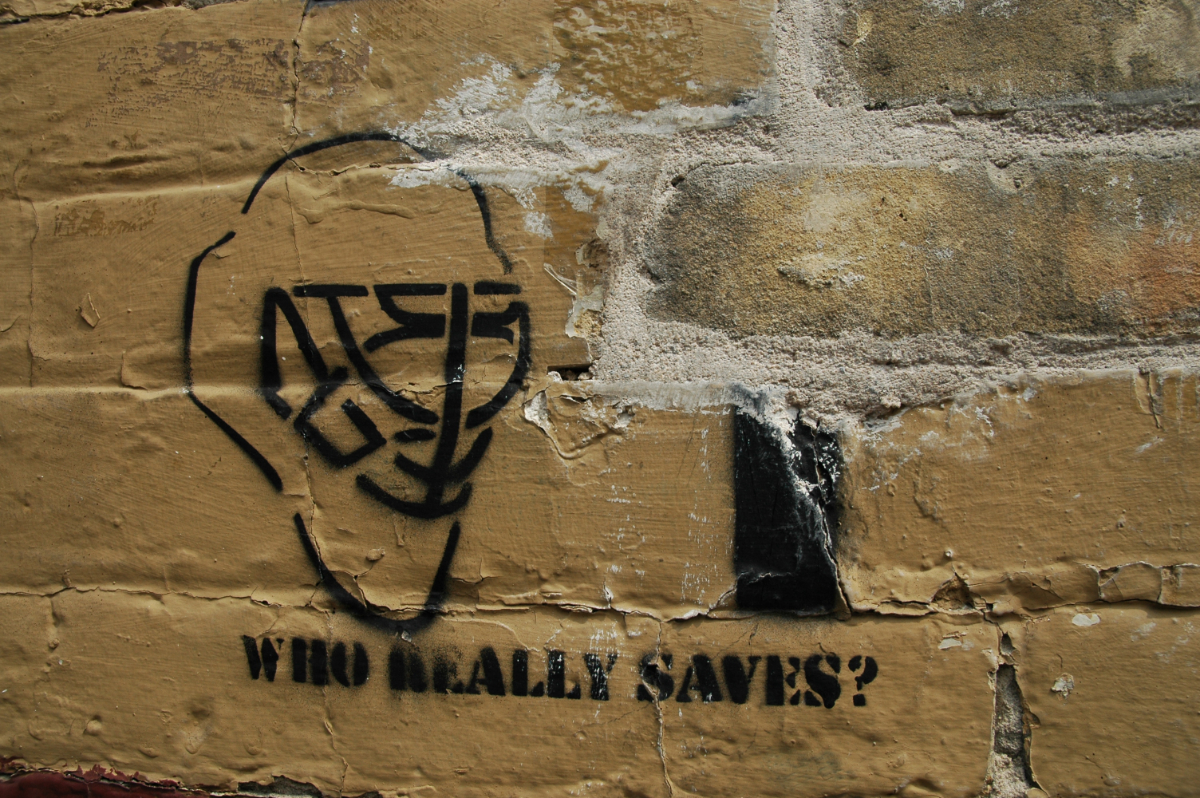 a stone wall painted brown with a stencil of a hockey goalie's mask and the caption 'who really saves?' spray painted in black