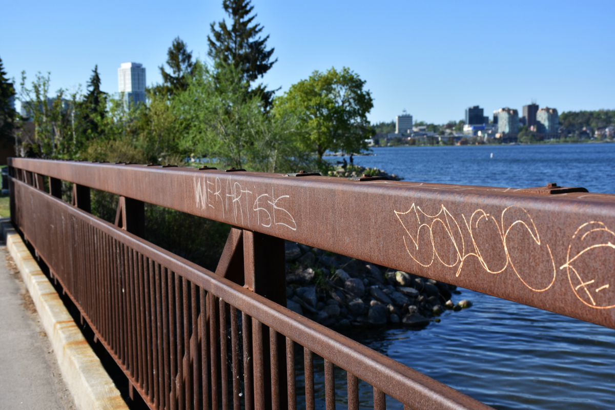 Barrie, ON A rusted metal guardrail on a bridge with a lake in the background. There is writing on the guardrail saying 'WE R ARTISTS' with less than ideal spacing.