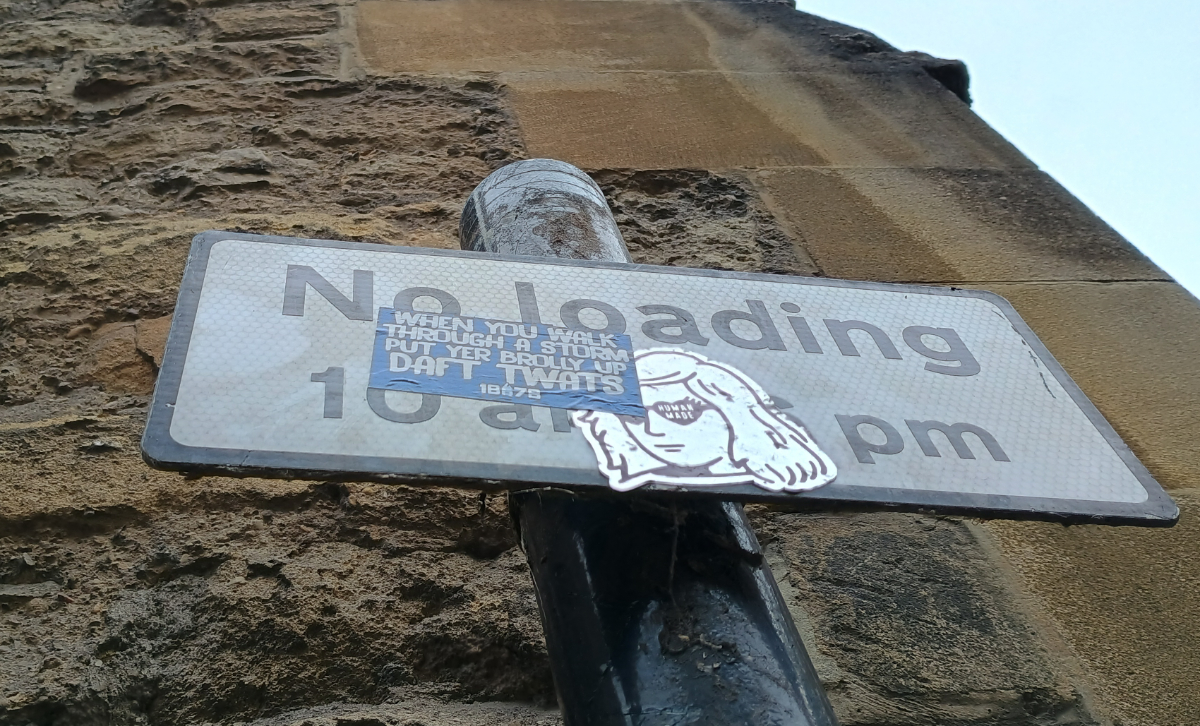 Oxford, UK A street sign in front of a decaying stone wall that says 'No loading 10am - 6pm' but part of it is covered with two stickers: a drawing of a woman wearing heart shaped sunglasses with 'human made' over one of the lenses, and a sticker saying 'when you walk through a storm put your brolly up DAFT TWATS'