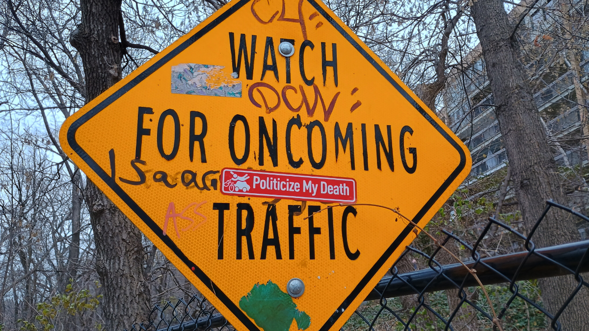 a diamond shaped sign saying 'WATCH FOR ONCOMING TRAFFIC' with various graffiti on it plus a sticker of a cyclist getting hit by a car and flying in the air with the text 'politicize my death'