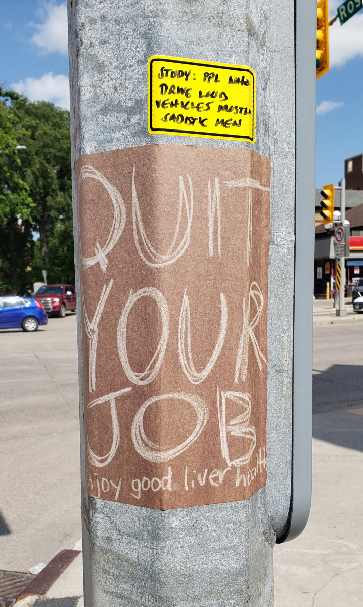 Silver lamp post with a brown paper sign saying 'QUIT YOUR JOB' in white writing on most of the 8.5x11 page, but small writing at the bottom that you can't entirely see, but it's enough to tell it says 'enjoy good liver health'