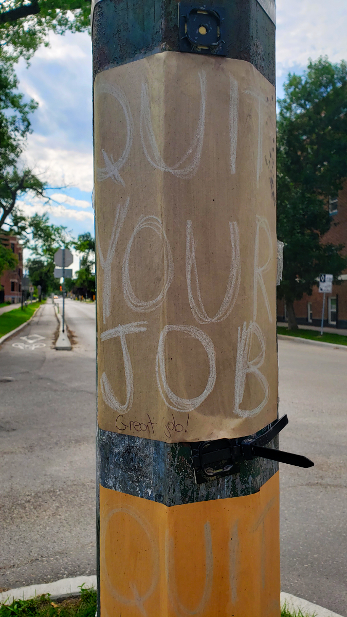 A brown paper QUIT YOUR JOB sign with white lettering on a green lamppost with 'Great job!' written at the bottom. There's another QUIT YOUR JOB poster below it but you can only see the QUIT part.