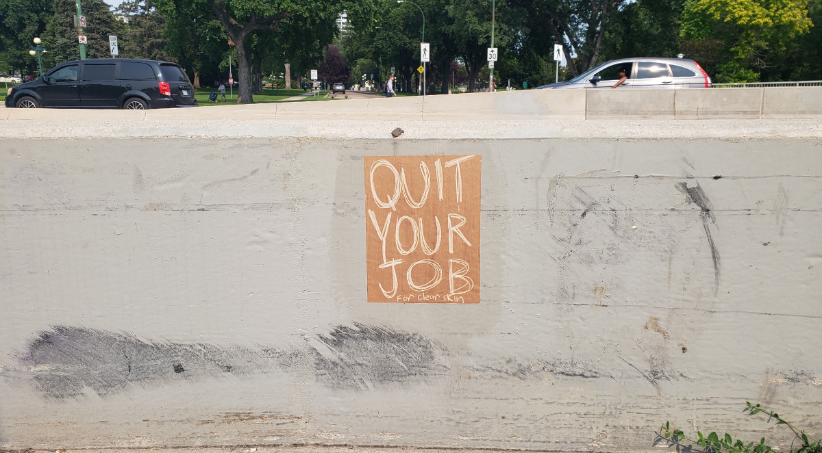 A concrete barrier between a sidewalk and a road with a QUIT YOUR JOB poster on brown paper with beige writing and the message 'For clear skin' at the bottom