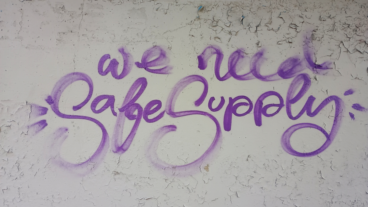 a concrete wall with whimsical writing in purple spray paint saying 'we need Safe Supply'