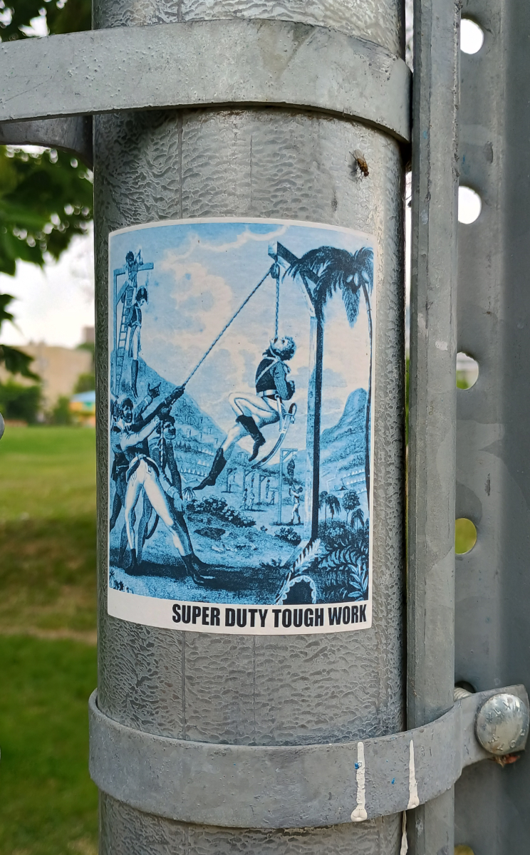 a metal pole with a poster featuring a painting of Hatians hanging their colonizers with a caption at the bottom saying 'super duty tough work'