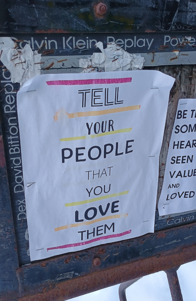 a community poster space on a bike rack with a 8.5x11 sheet with various fonts and multiple colours of underline saying 'tell your people that you love them'