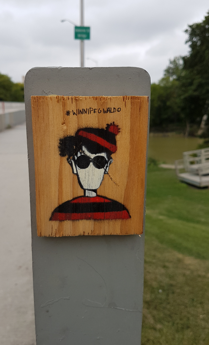 The end of a bridge railing with a wooden block glued to it that has an expressionless image of a darker version of Waldo/Wally with red and black garb instead of red and white, and the caption at the top #winnipegWaldo