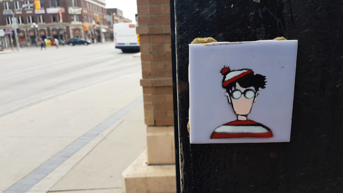 A light blue tile on a black lamp post with an expressionless face of Waldo/Wally with the normal red and white colours. There is a blurry street in the background.