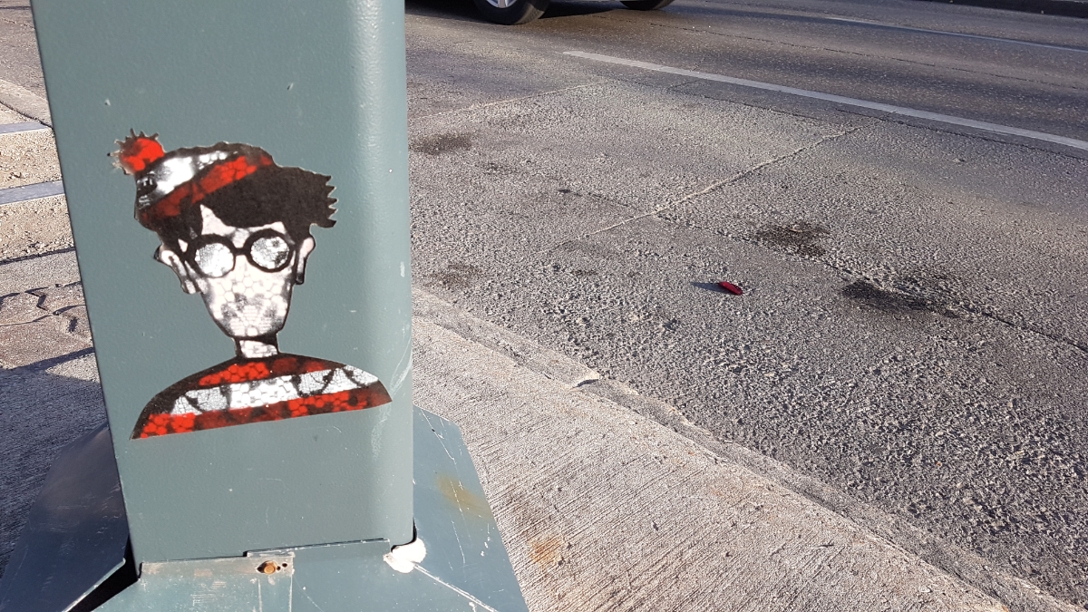 A thick grey trunk of a parking ticket station with a sticker on it of an expressionless Waldo/Wally with the normal red and white colours and a sort of smudged black hexagon type pattern within it.