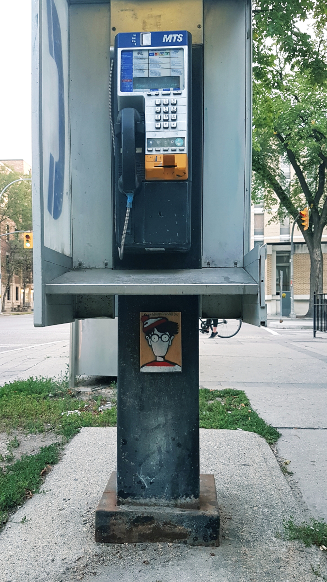 A phone booth with a wooden block with Waldo/Wally's face on it (no nose or mouth but at least there are pupils this time!) and the #winnipegWaldo hashtag.