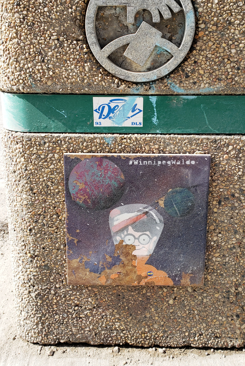 A street trash bin with a gravel/stone facade, with a purple tile with planets and stars in the background, and Waldo/Wally in an orange spacesuit, smiling like he does in the books, and #WinnipegWaldo in a serif typeface at the top.