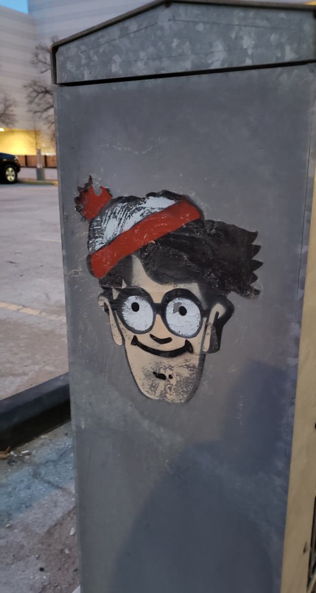 A grey metal box on the street with a sticker of a standard Waldo/Wally just looking up, smiling at us all.