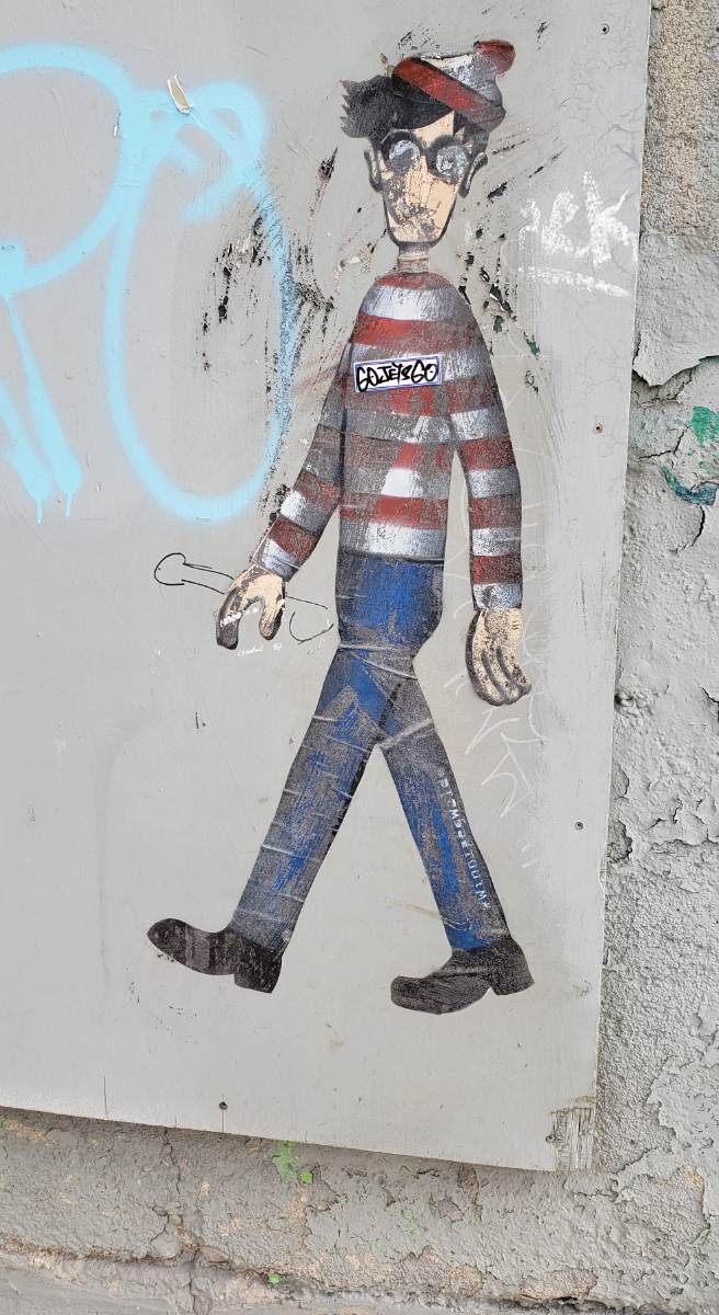 A full body walking expressionless Waldo/Wally painted on a grey piece of wood on a painted concrete wall. The painting is a bit worn for wear, with other gibberish tags painted around it and a giant dick painted protruding from more or less where Waldo's actual giant dick would come from.