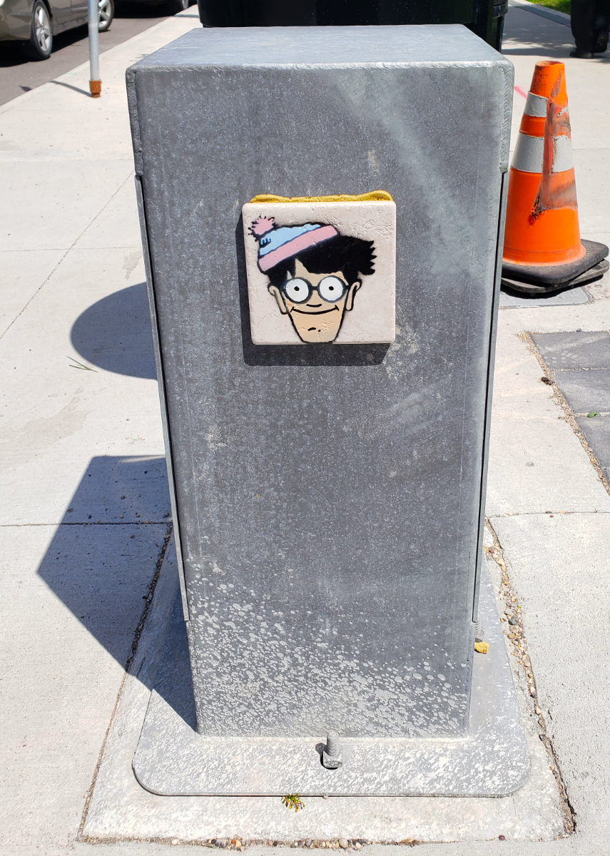 A dull grey box on a sidewalk with light pinky beige tile glued to it featuring a smiling Waldo/Wally wearing a hat in light blue and light pink, because fuck yeah trans rights!