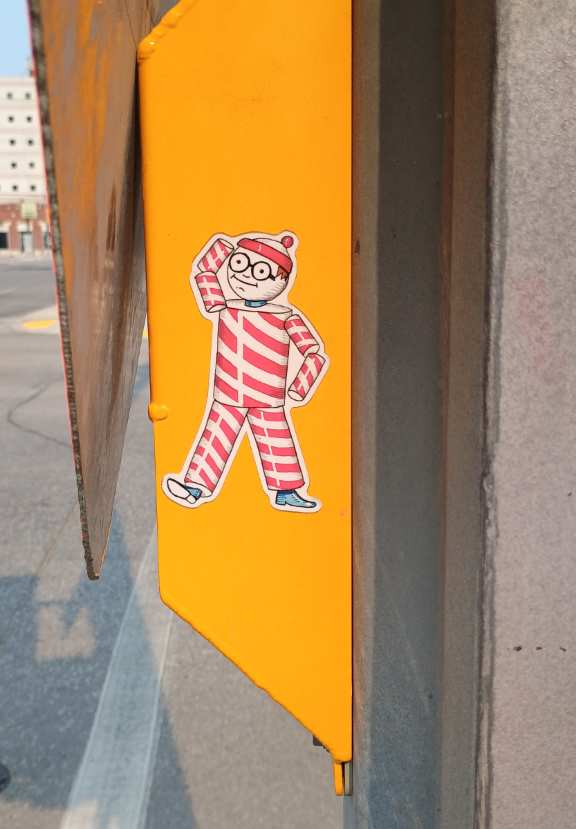 A yellow crosswalk speaker with a sticker of the Nutty Club candy brand mascot, RIP Winnipeg legend, with a Waldo-like face on the round head