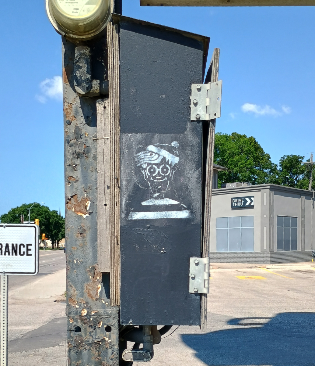 A black box on a metal pole with Winnipeg Waldo spray painted in white from a stencil. In the background is a street to the left and a Starbucks drive-thru to the right.