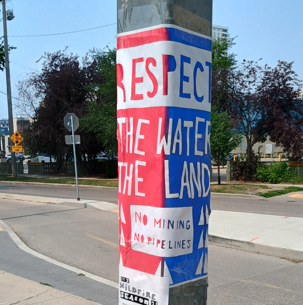 a half red and half blue background sign pasted to a square pole, with a white box at the top with letters cut out to say 'respect' exposing the split background, and text below saying 'the water' and 'the land' with white triangles representing trees surrounding another box at the bottom saying 'no mining, no pipelines'