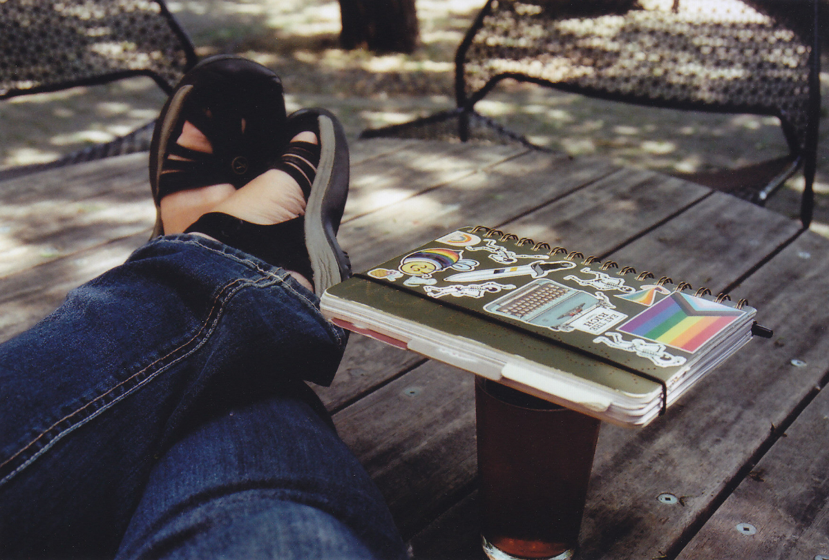 sandaled feet resting upon a wooden table outdoors with a notebook covered in queer and/or leftist stickers placed on top of a glass of beer