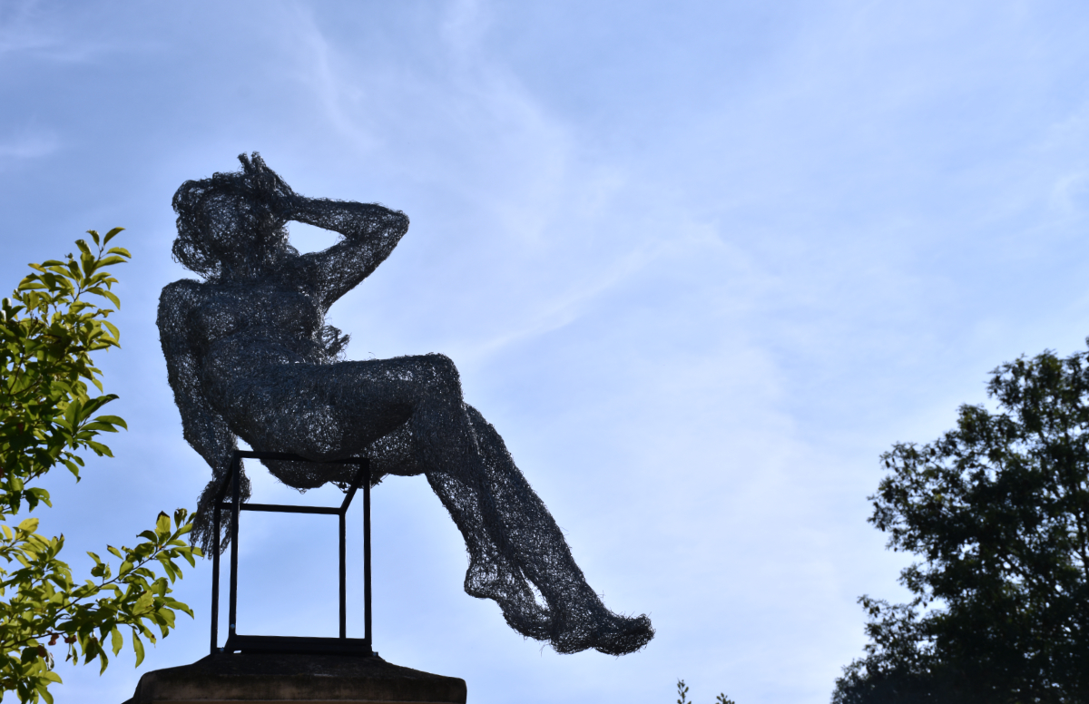 the silhouette of a sculpture made of wires that looks like a vaguely feminine body sitting on a cube and leaning back with the back of her hand on her forehead against a mostly blue sky with some green foliage sticking out from just outside the frame.
