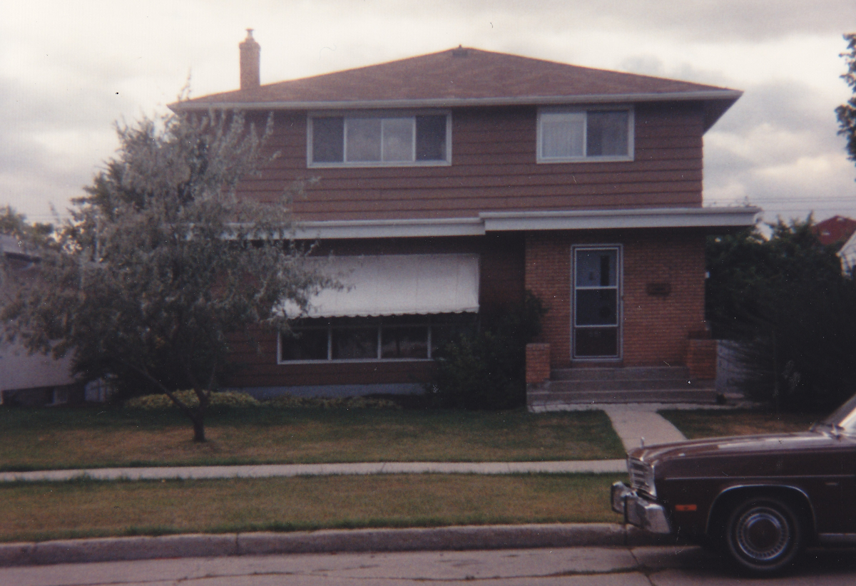 A brown two-storey house with a foundation and steps up to a door on the right side, with a large living room window to the left and two bedroom windows on the second floor. A 70s era car hood is visible on the street in front of the house. There is a tree in the front yard that is well-mowed.